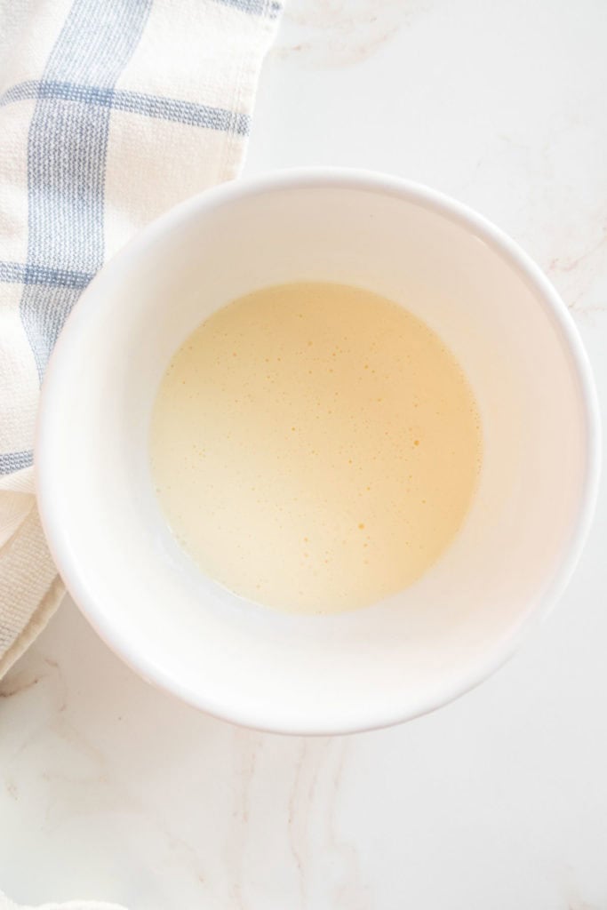 A white bowl filled with a light beige liquid, placed next to a white and blue checkered cloth on a marble surface.