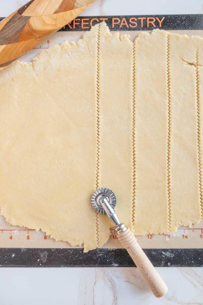 A rolled-out sheet of pastry dough with evenly spaced cuts made by a pastry wheel, lying on a gridded pastry mat with a wooden rolling pin partially visible at the top.