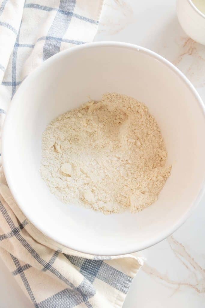 A white bowl contains a beige powdery substance, likely flour, on a countertop with a checkered cloth beside it.