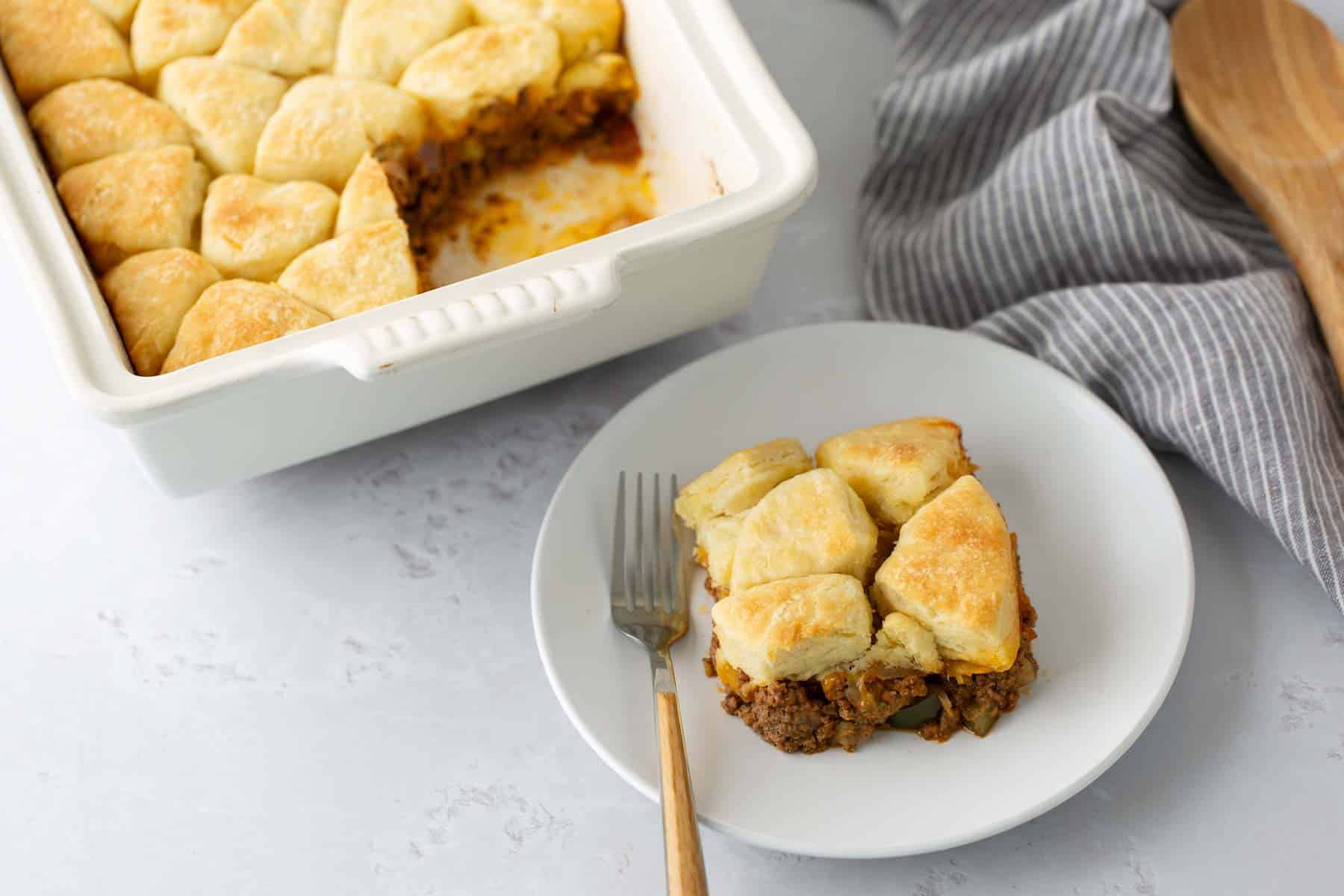 A baking dish of biscuit-topped ground meat casserole with one serving on a white plate, accompanied by a fork. A gray striped napkin and a wooden spoon are beside the dish.