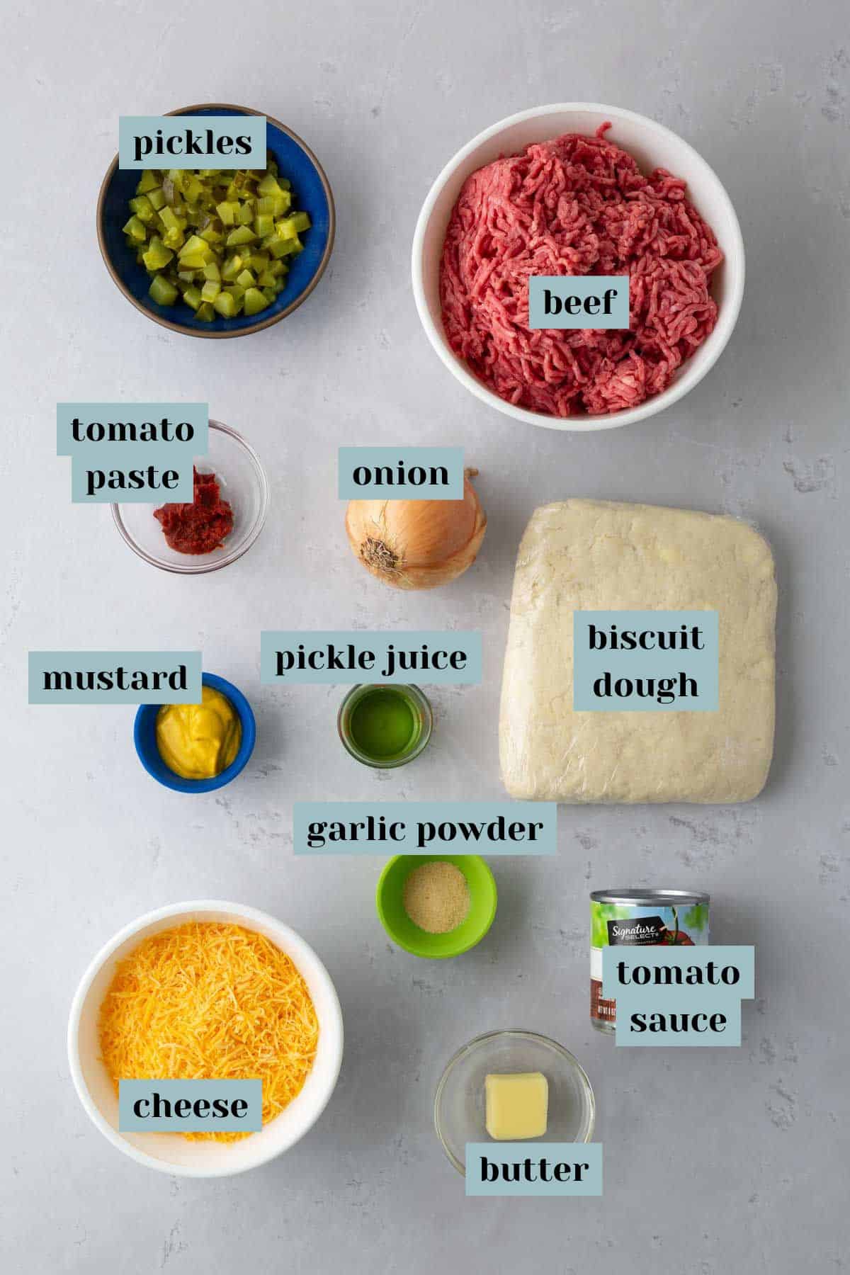 Ingredients arranged on a grey surface including pickles, ground beef, biscuit dough, shredded cheese, mustard, tomato paste, tomato sauce, pickle juice, onion, garlic powder, and butter.