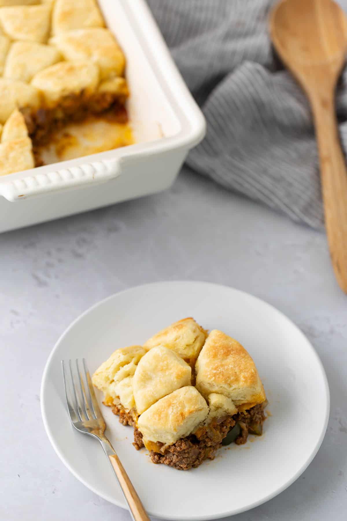 A white plate holds a serving of beef casserole with biscuit topping. A white baking dish with the remaining casserole is in the background, alongside a wooden spoon and a striped cloth.