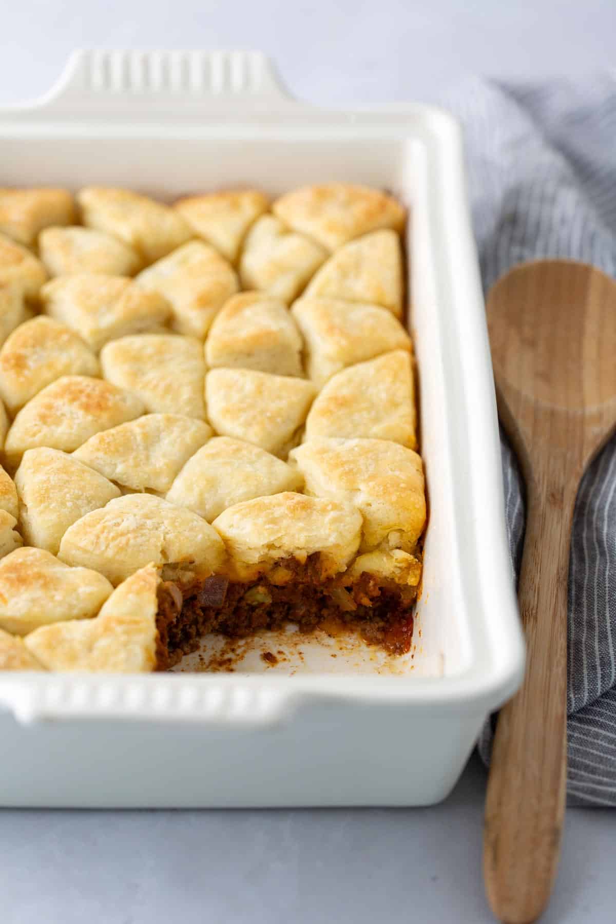 A white baking dish with a golden-brown, biscuit-topped casserole. A portion has been served, revealing a savory filling beneath. A wooden spoon rests next to the dish on a striped cloth.