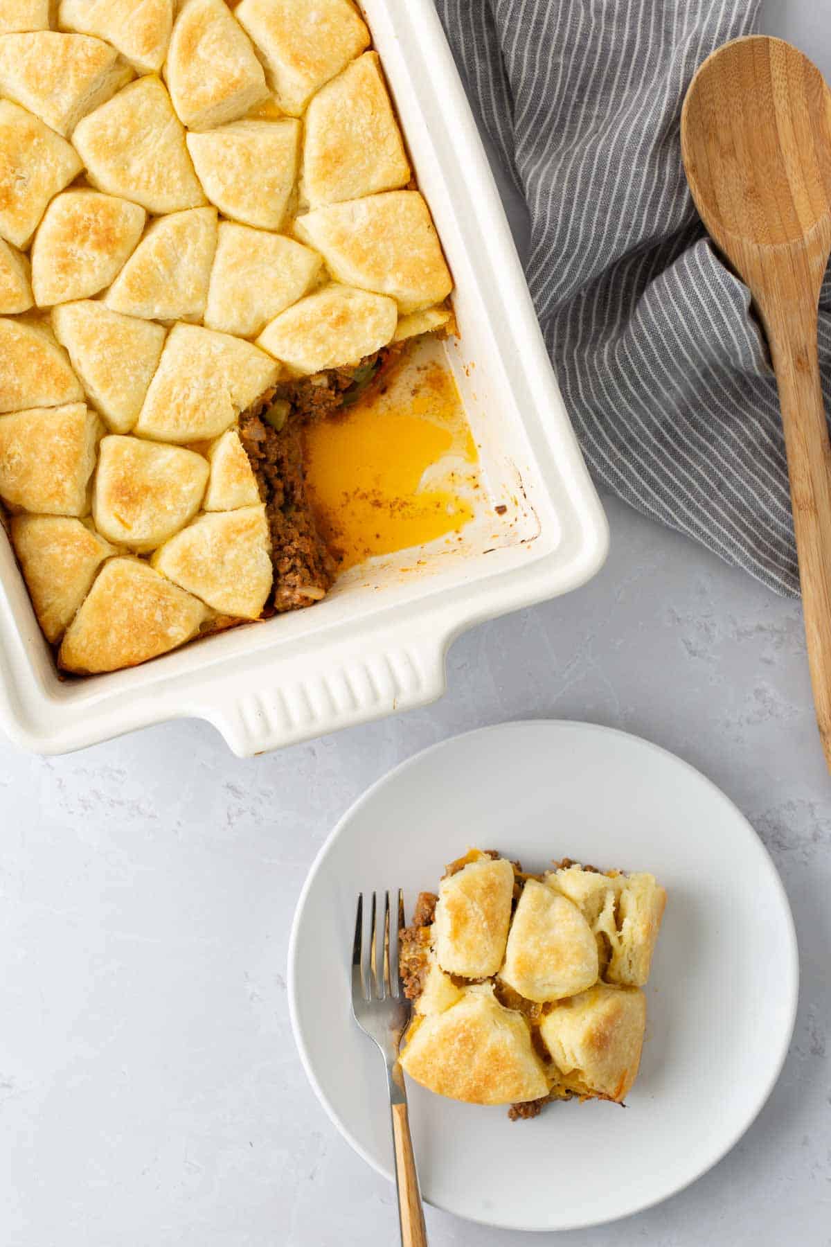 A casserole dish filled with a baked, golden brown biscuit-topped casserole. One serving is placed on a white plate with a fork. A striped cloth and wooden spoon are beside the dish.