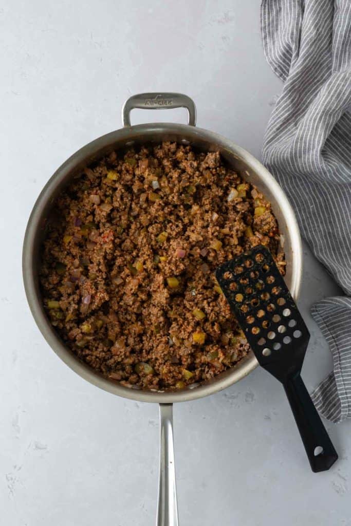 A pan filled with cooked ground meat mixed with diced vegetables. A black spatula sits on the edge of the pan, with a gray striped towel beside it.