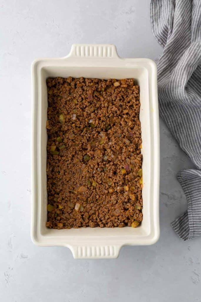 A rectangular baking dish filled with seasoned ground beef is placed on a light gray surface next to a gray and white striped cloth.