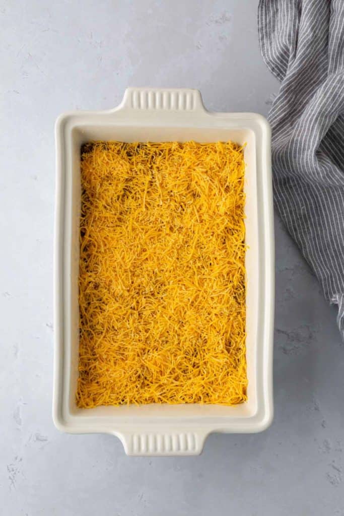 A rectangular white baking dish filled with shredded yellow cheese, placed on a light gray surface next to a gray striped cloth.