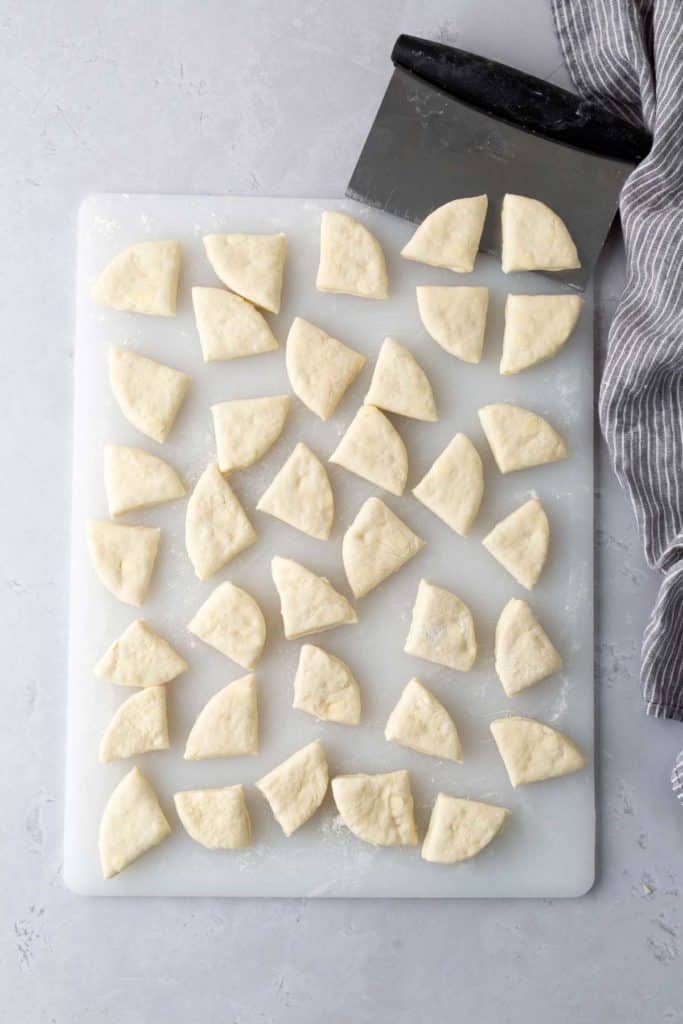 A cutting board with numerous semi-circular pieces of dough, a dough scraper, and a striped cloth on the side.