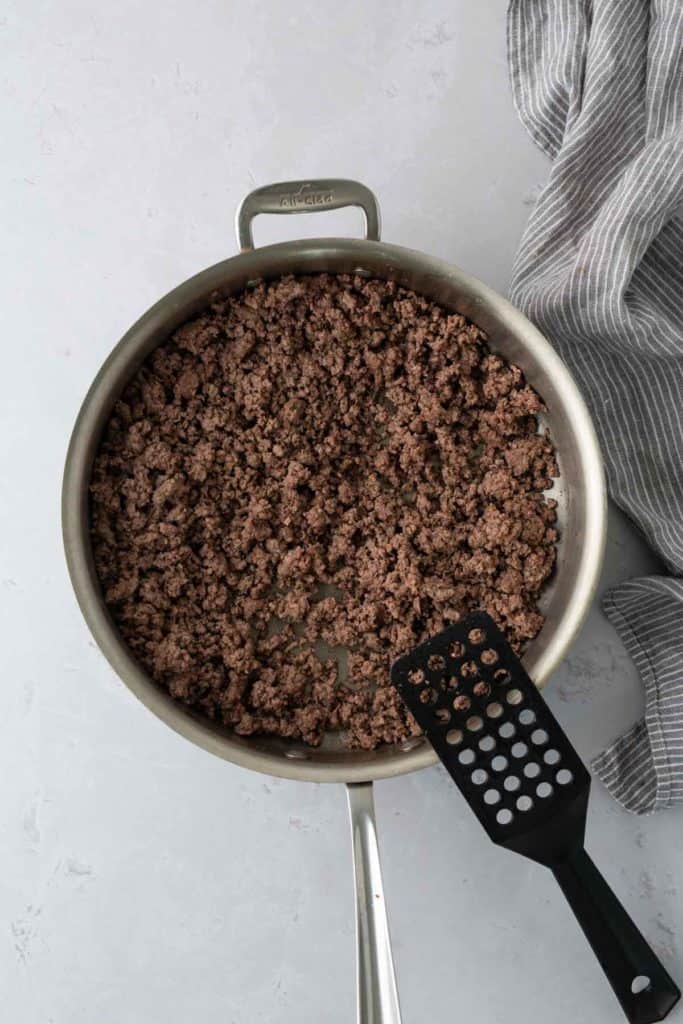 A skillet containing cooked ground beef with a black spatula next to a gray-striped cloth.