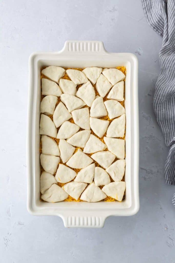 A rectangular casserole dish filled with uncooked, triangular dough pieces arranged on top of a layer of shredded cheese.