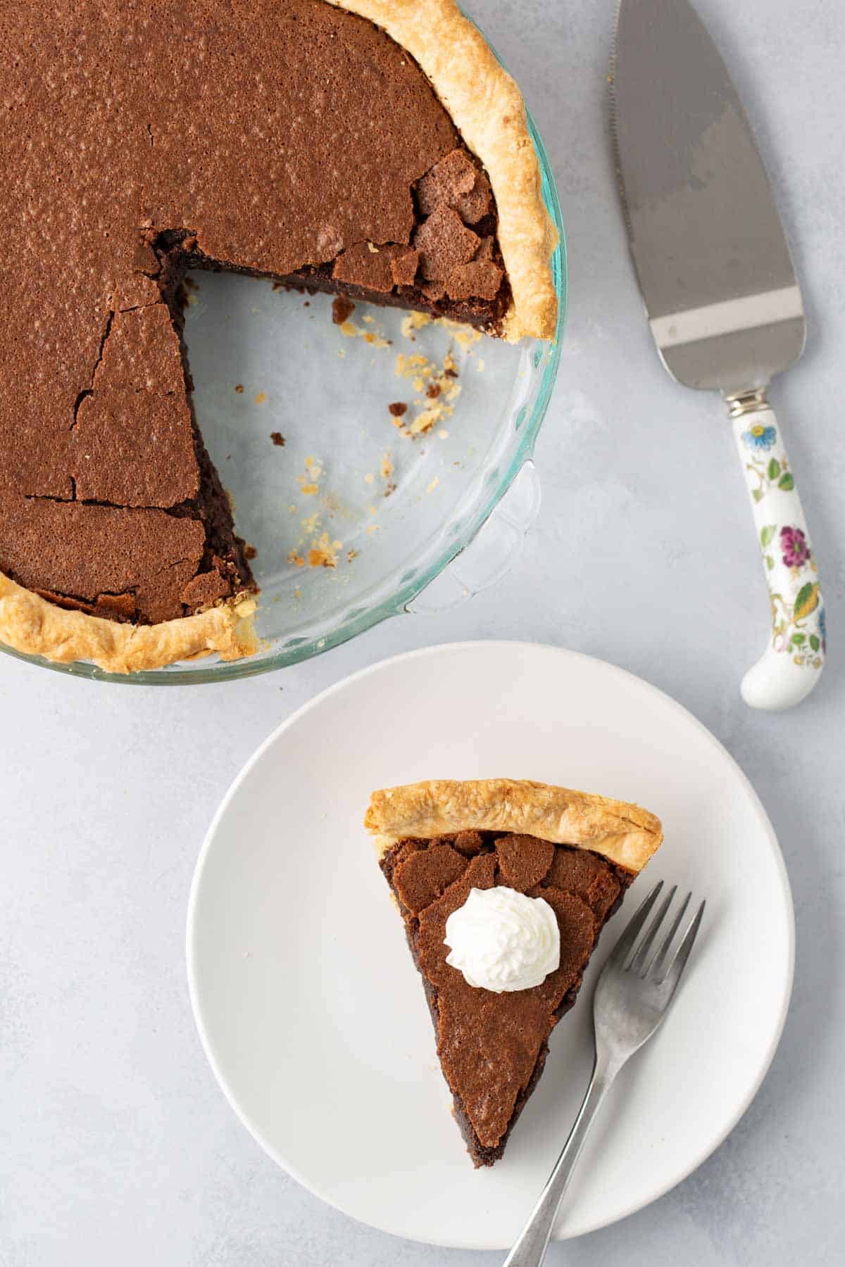 A pie with a missing slice is next to a dessert plate. The plate has a single slice of pie topped with whipped cream, a fork, and a pie server with a floral handle on a light gray surface.