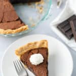 A slice of chocolate chess pie topped with whipped cream on a white plate with a fork beside it. The rest of the pie and pieces of dark chocolate are visible in the background.