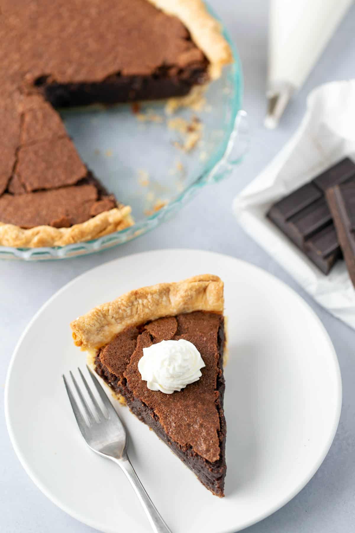 A slice of chocolate chess pie topped with whipped cream on a white plate with a fork beside it. The rest of the pie and pieces of dark chocolate are visible in the background.