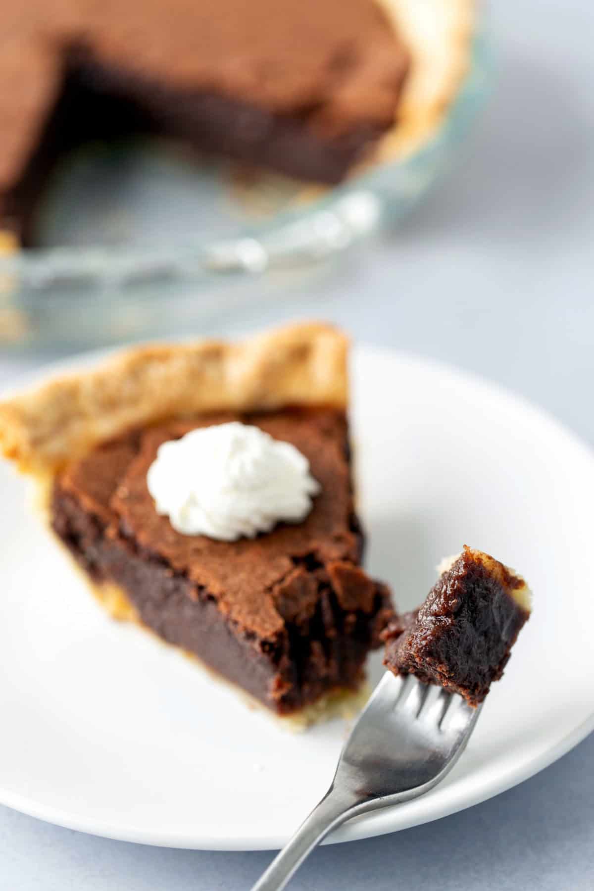 A slice of chocolate pie with a dollop of whipped cream on a white plate, with a fork holding a bite-sized piece of the pie.