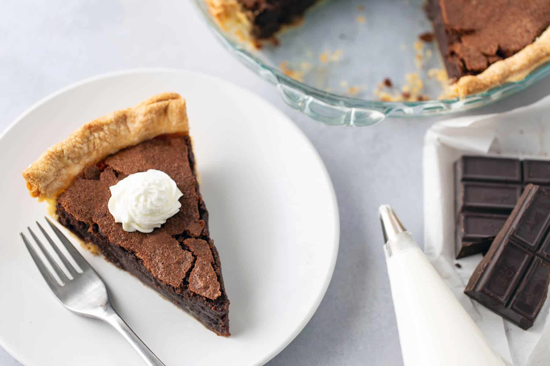 A slice of chocolate pie with a dollop of whipped cream on a white plate, accompanied by a fork. The pie dish with remaining pie, chocolate pieces, and a piping bag are in the background.