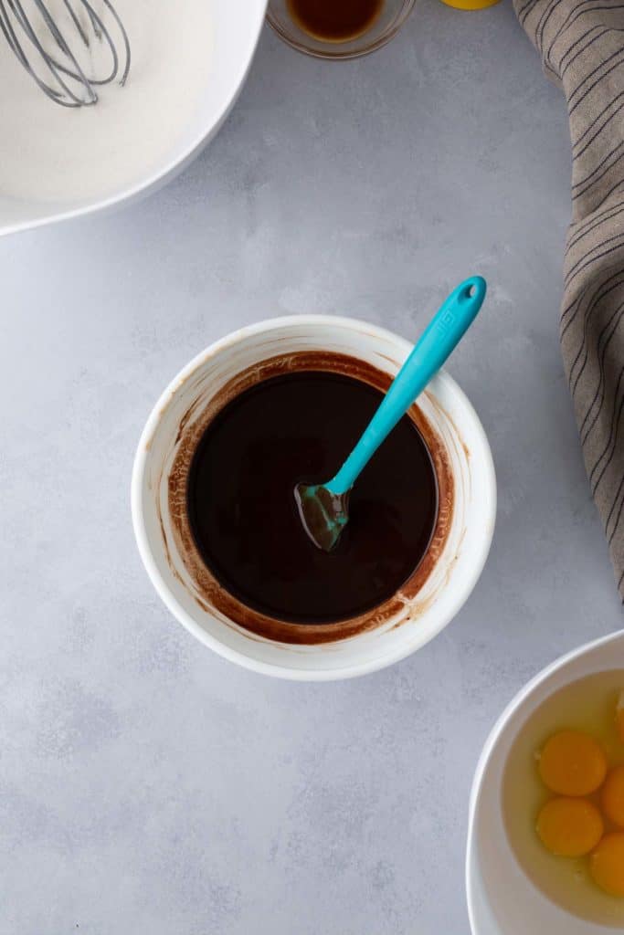 A mixing bowl with a blue-handled spatula sits on a gray surface, containing a dark mixture, likely chocolate. Nearby are bowls with sugar, vanilla extract, and eggs.