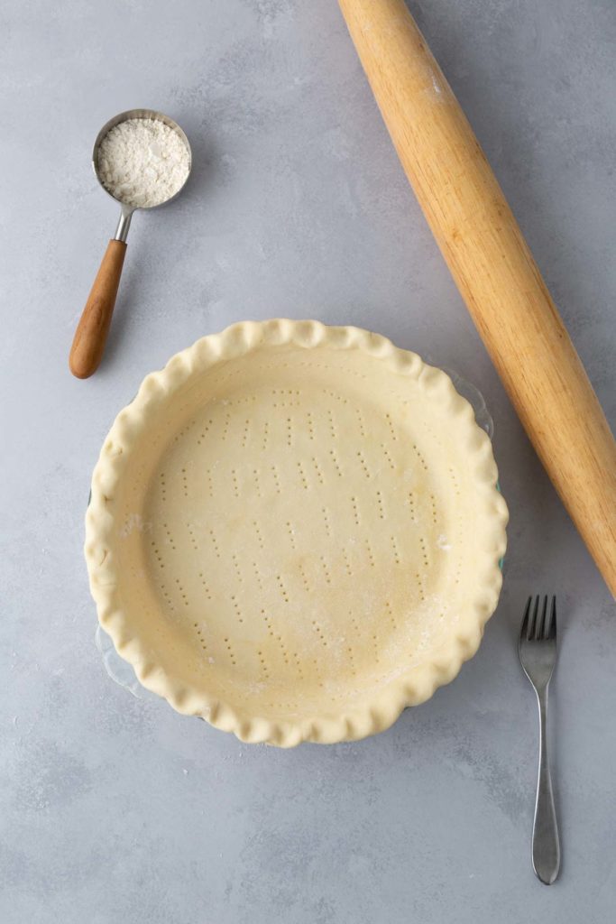 A prepared pie crust in a fluted pie dish with a fork, rolling pin, and a measuring cup of flour placed on a gray surface.