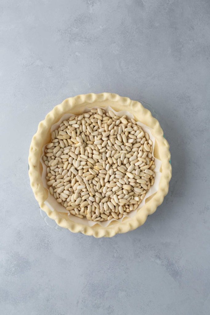 Unbaked pie crust filled with dried beans, used as pie weights, set on a gray surface.