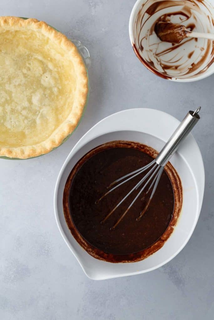 Top-down view of an unbaked pie crust in a dish, a bowl with a whisk inserted into chocolate filling, and a bowl with remnants of chocolate.