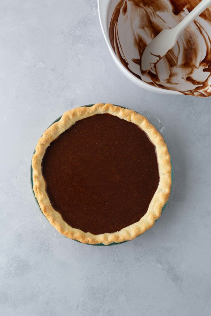 A freshly baked chocolate pie is in the foreground with a pie crust and a smooth chocolate filling. In the background, a mixing bowl with chocolate remnants and a spatula is visible.