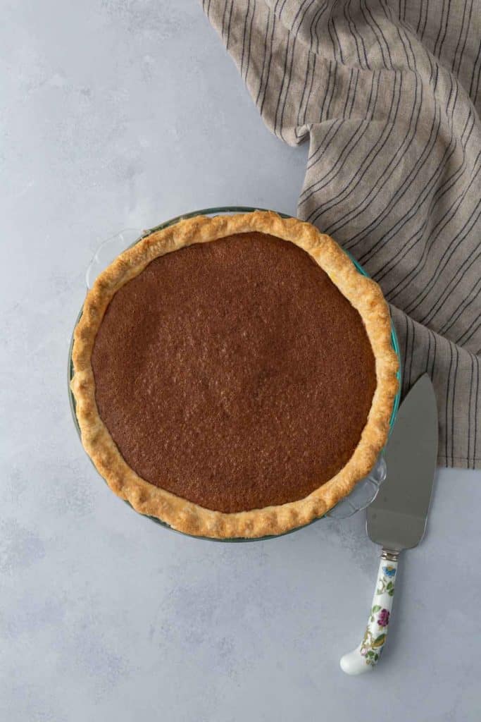 A whole pie with a browned top in a glass pie dish on a light gray surface. A knife with a floral handle and a striped cloth are placed beside it.