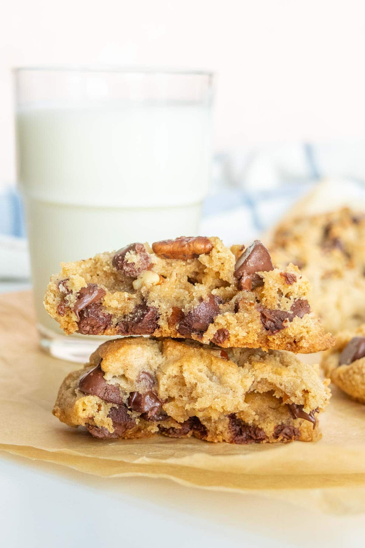 A glass of milk is placed behind two stacked chocolate chip cookies on a piece of parchment paper.