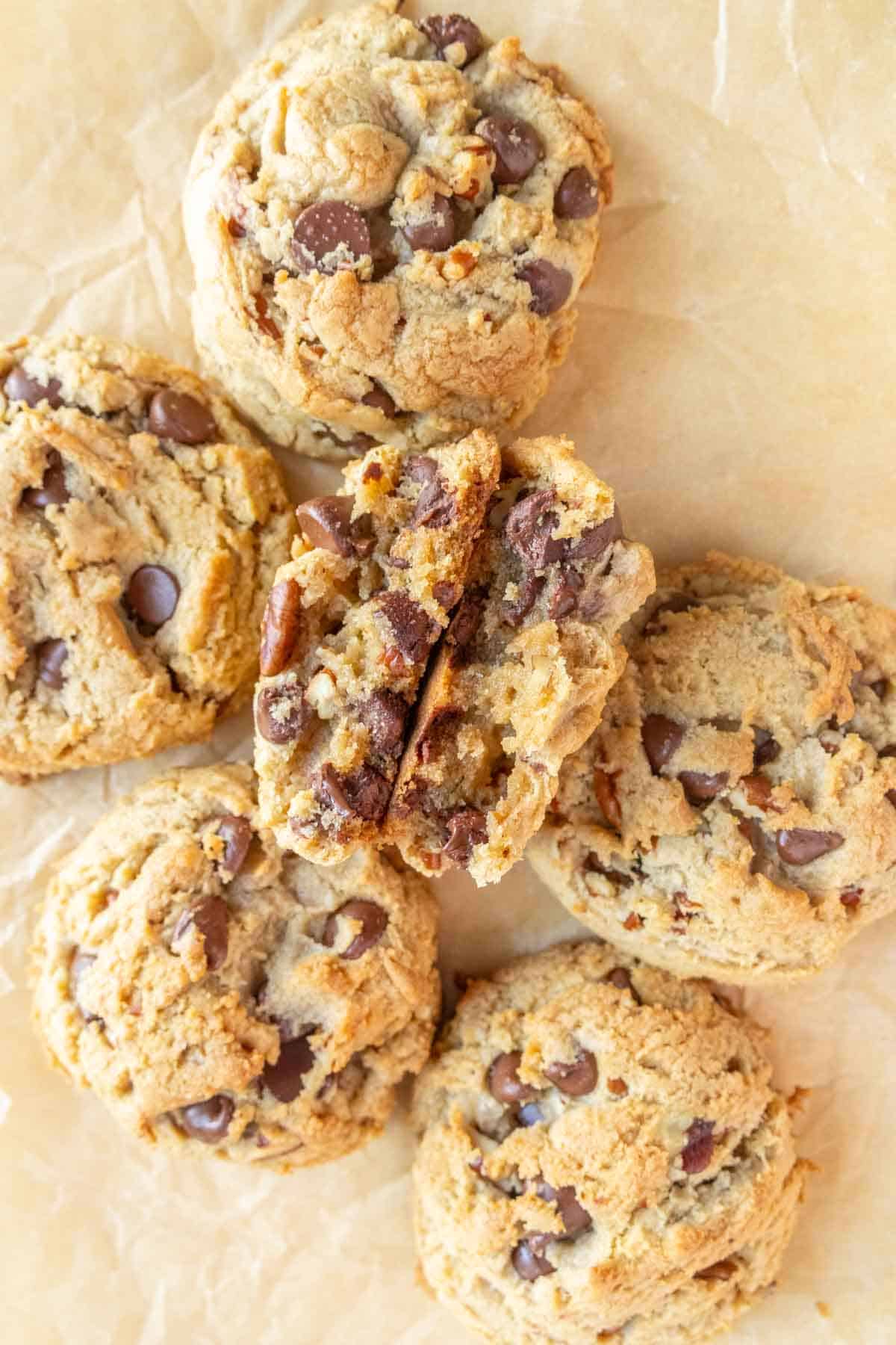 Six chocolate chip cookies, one broken in half, displayed on a sheet of parchment paper.