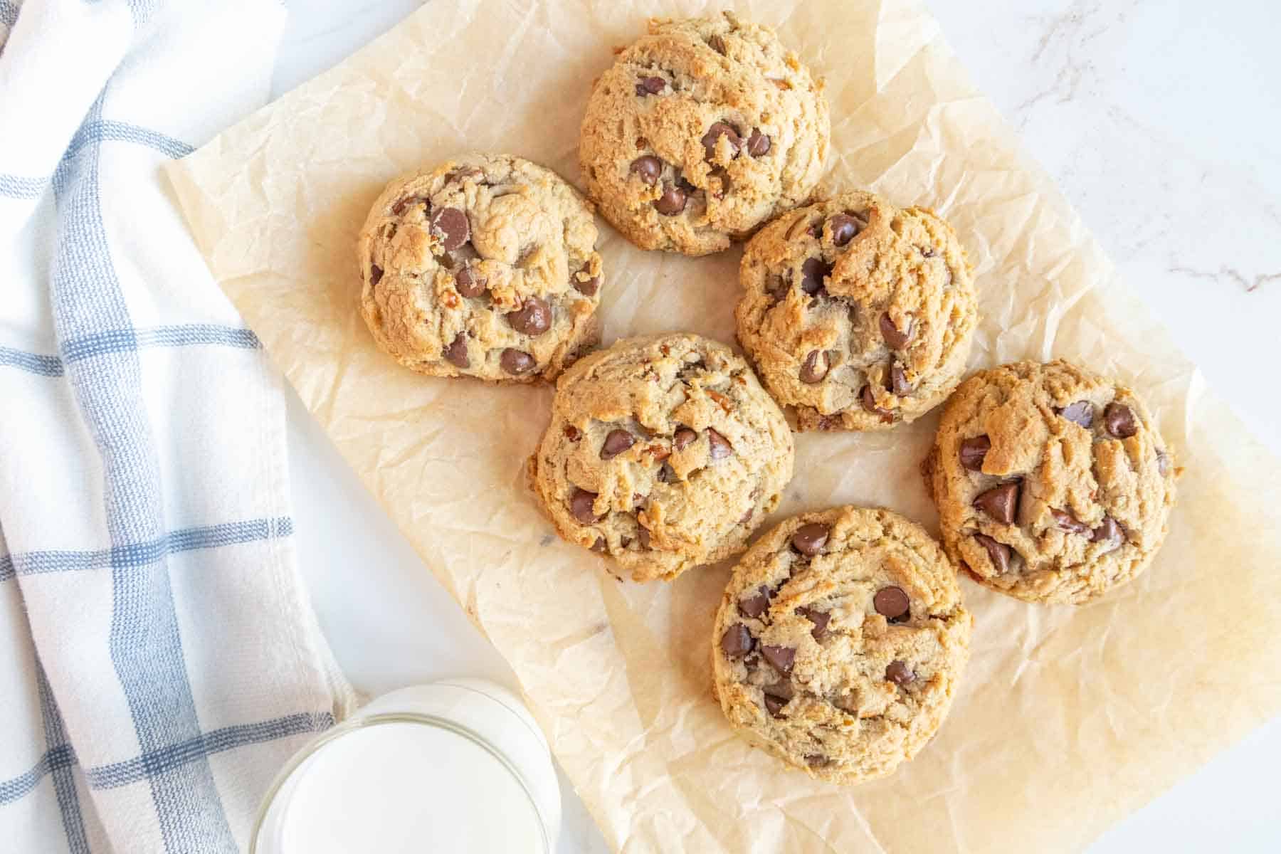 Freshly baked chocolate chip cookies sit on parchment paper next to a glass of milk and a blue-checked cloth.