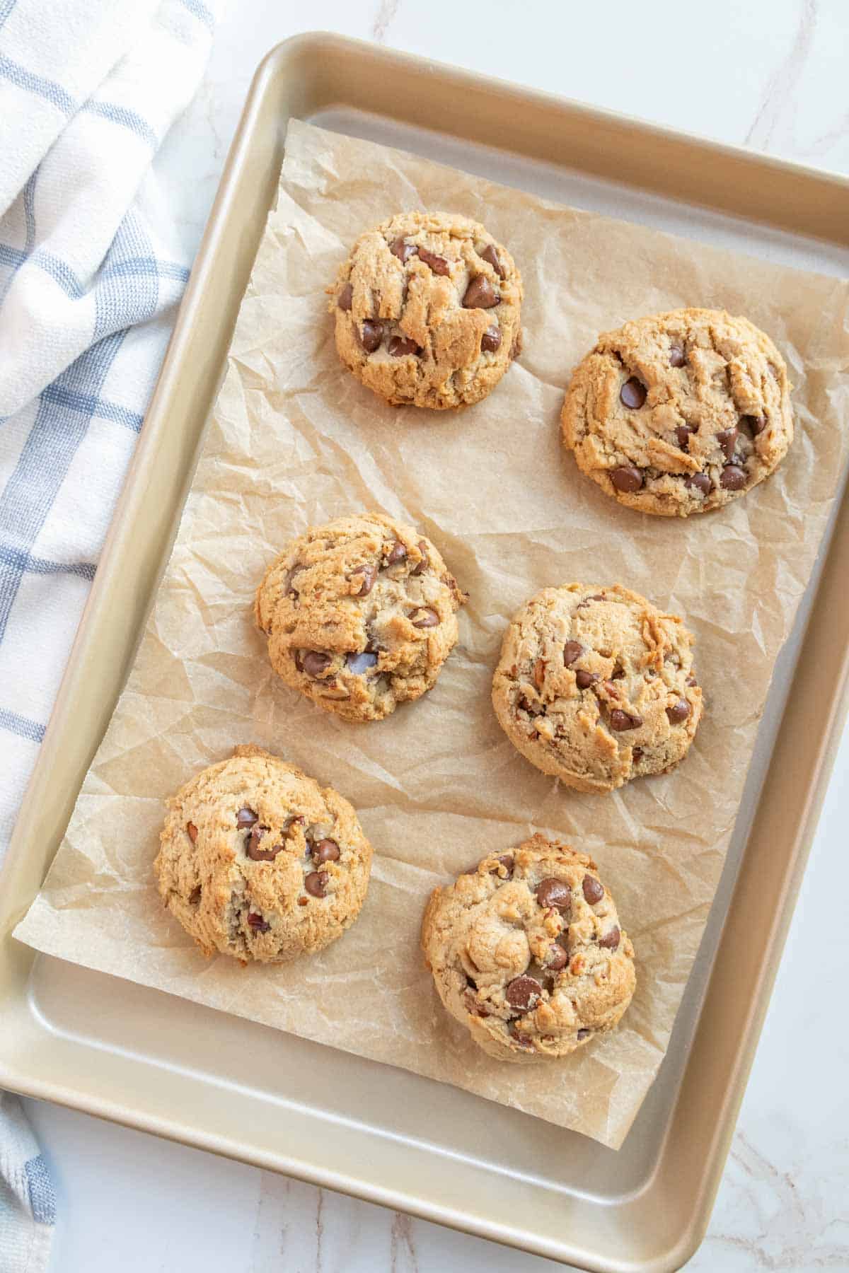 A baking tray with six freshly baked chocolate chip cookies on parchment paper, next to a folded striped kitchen towel.