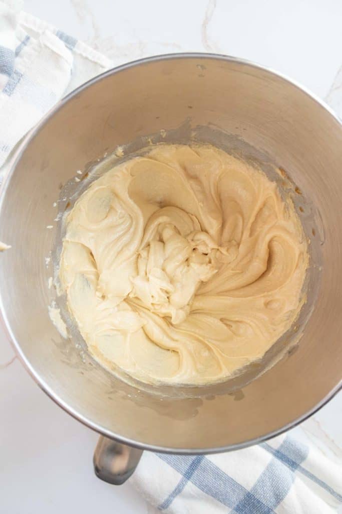 A stainless steel mixing bowl contains creamy, light-colored batter. A blue and white checkered cloth is partially visible underneath.