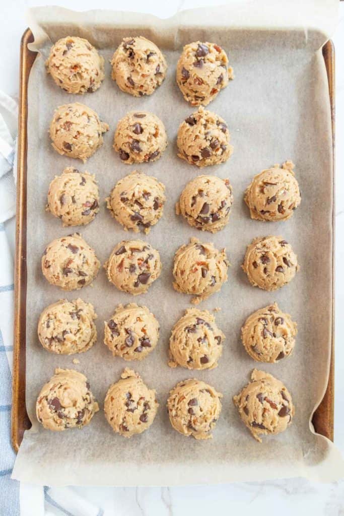 A baking tray lined with parchment paper holds 24 raw cookie dough balls, each containing chocolate chips, arranged in a 4x6 grid pattern.