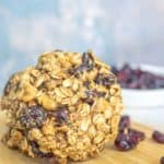 A large oatmeal and dried fruit cookie is placed on a wooden surface. Dried cranberries are scattered around, with a white bowl filled with more dried cranberries in the background.