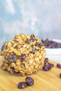 A large oatmeal and dried fruit cookie is placed on a wooden surface. Dried cranberries are scattered around, with a white bowl filled with more dried cranberries in the background.