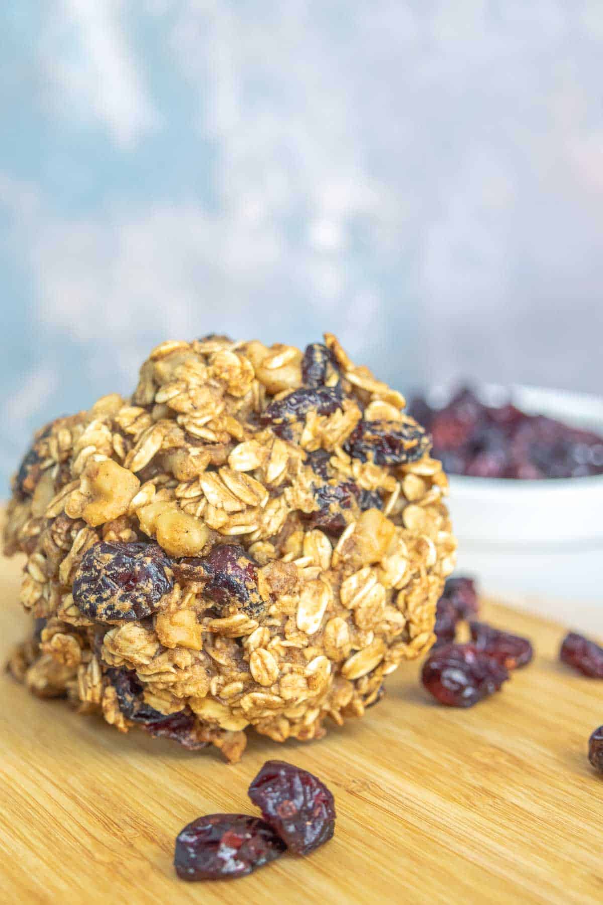 A large oatmeal and dried fruit cookie is placed on a wooden surface. Dried cranberries are scattered around, with a white bowl filled with more dried cranberries in the background.