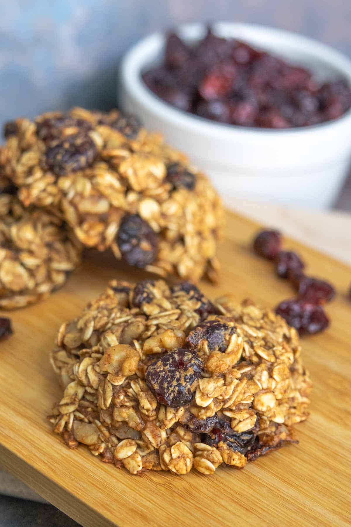 Close-up of oat and dried fruit cookies on a wooden board, with a white bowl of dried fruit in the background.