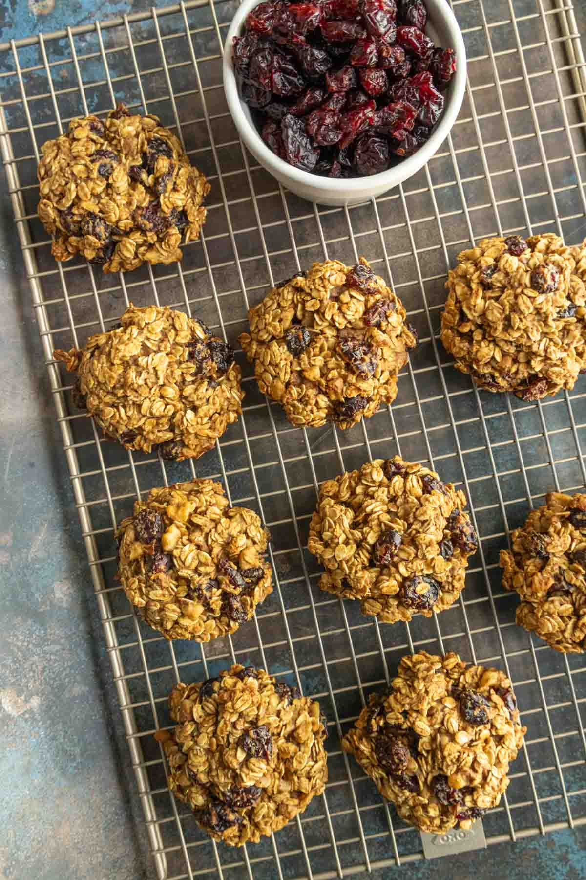 Nine oatmeal cookies with dried fruit are arranged on a cooling rack, with a bowl of additional dried fruit placed above them.