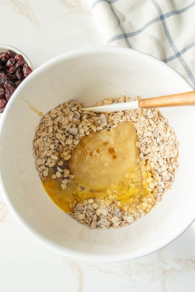 A mixing bowl with rolled oats, applesauce, egg, and a spoon ready for mixing. A small bowl of cranberries is partially visible at the top left, and a cloth is partly seen on the right.