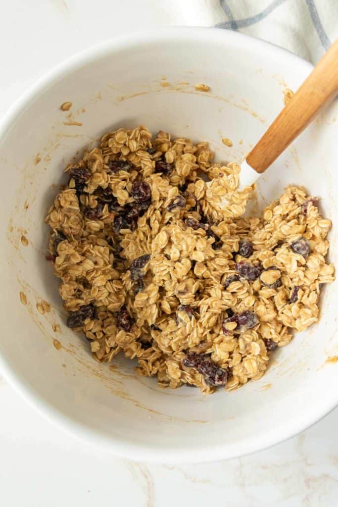 A mixing bowl containing a blend of oats, raisins, and a creamy ingredient, being stirred with a wooden spoon.