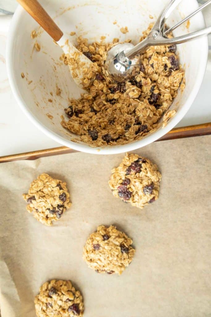 A mixing bowl containing oatmeal mixture with raisins and a metal scoop. Some scooped oatmeal portions are placed on a parchment-lined baking tray.