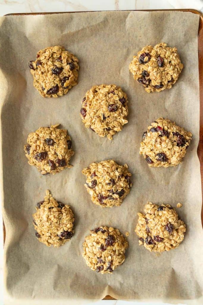 A baking tray lined with parchment paper holds eight freshly baked oatmeal cookies with visible raisins.