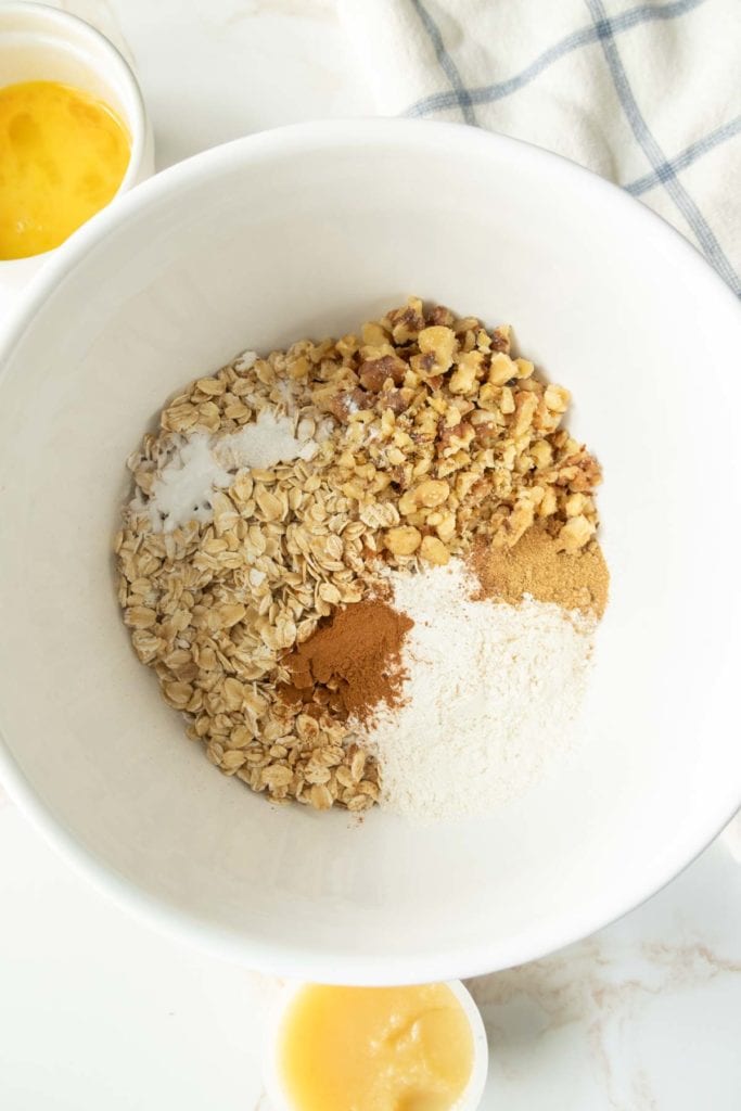 A mixing bowl containing oats, chopped nuts, flour, cinnamon, and a blend of other dry ingredients. A cup of beaten egg and a cup of applesauce are nearby.