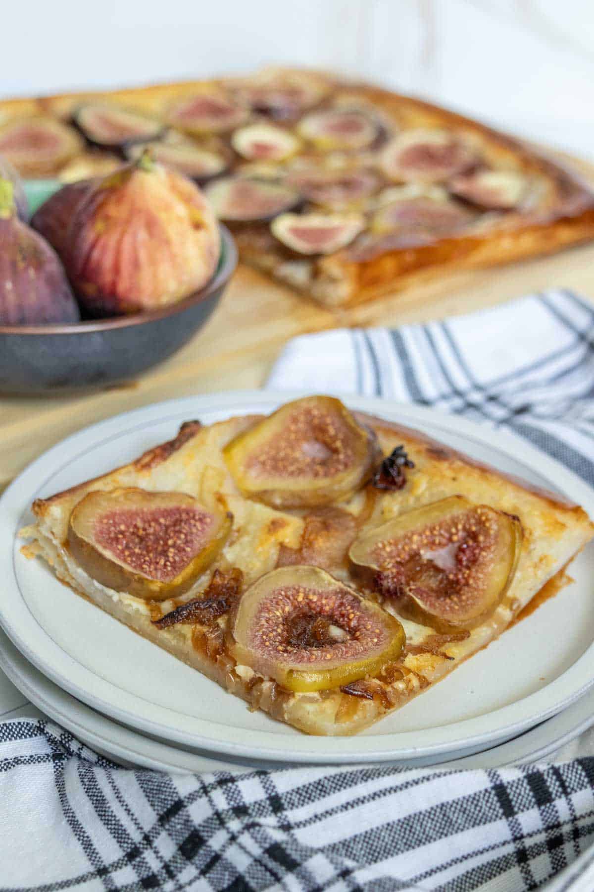 A slice of fig tart on a white plate with the whole tart and fresh figs in a bowl in the background.