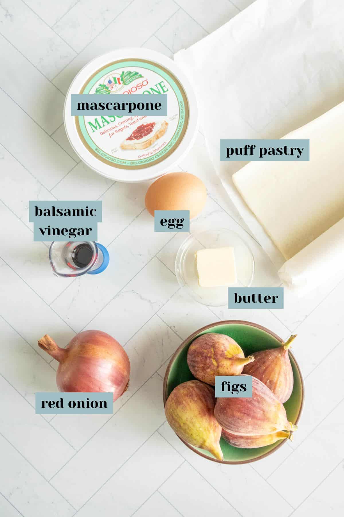 Ingredients for a recipe are laid out, including mascarpone, puff pastry, balsamic vinegar, an egg, butter, red onion, and figs.