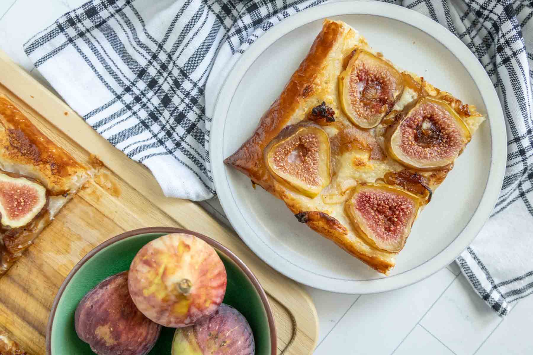 A plate with a single slice of fig tart is placed next to a wooden board with more tart slices and a bowl containing whole figs. A striped kitchen towel is partially visible in the background.