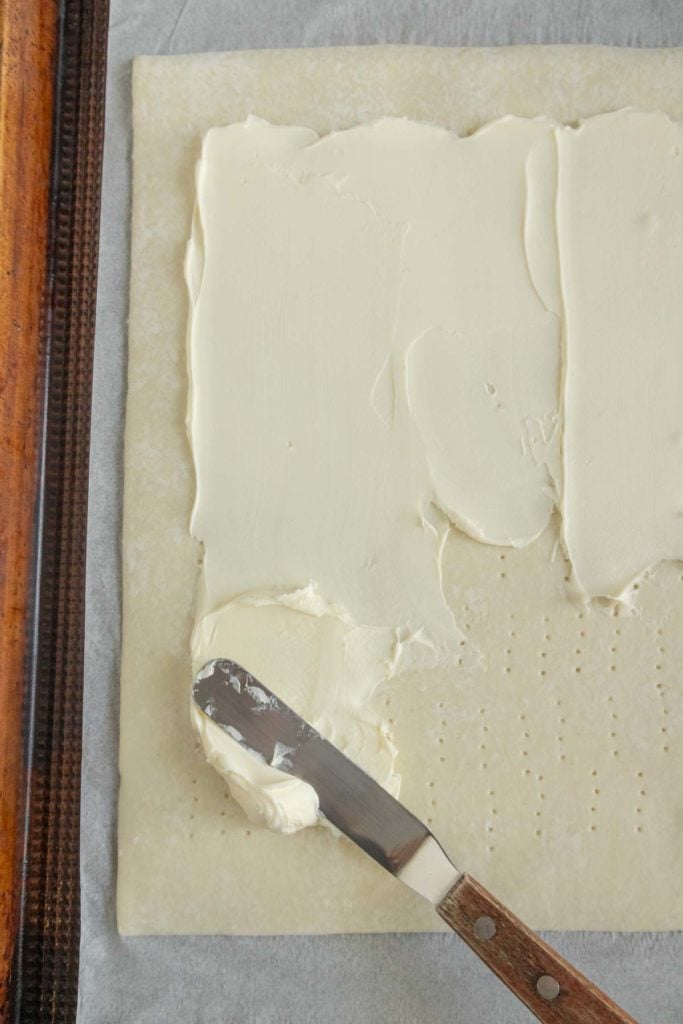 A metal spatula spreads a layer of cream cheese onto a rectangular sheet of pastry dough, which has been pricked with small holes.