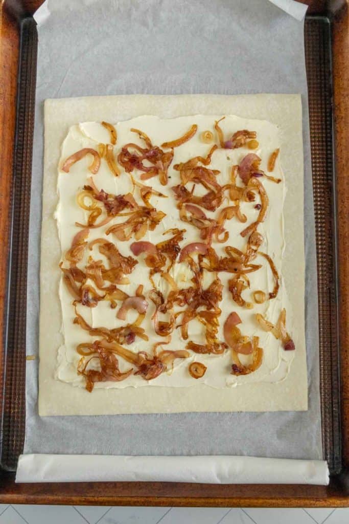 Rectangular pastry sheet topped with caramelized onions and cream cheese on a baking tray lined with parchment paper.