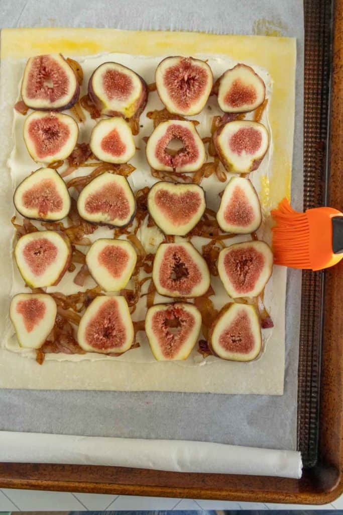 A baking tray with a partially assembled puff pastry topped with sliced figs and caramelized onions. An orange brush is applying egg wash to the pastry edges.