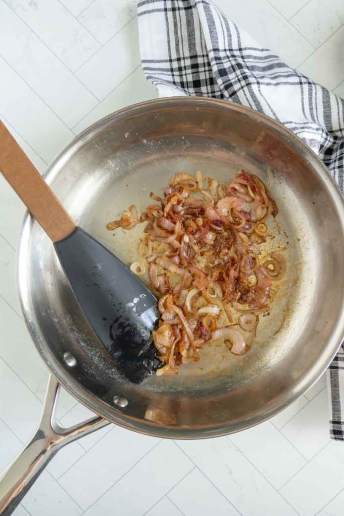 Sautéed onions and garlic are being cooked in a silver frying pan using a black spatula, with a black and white checkered cloth nearby.