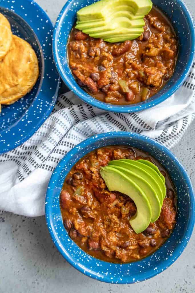 Two blue bowls of chili topped with sliced avocado are set on a table with a white and black striped cloth. A plate of biscuits is visible beside the bowls.