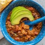 A blue bowl filled with chili garnished with avocado slices and served with a biscuit on the side.